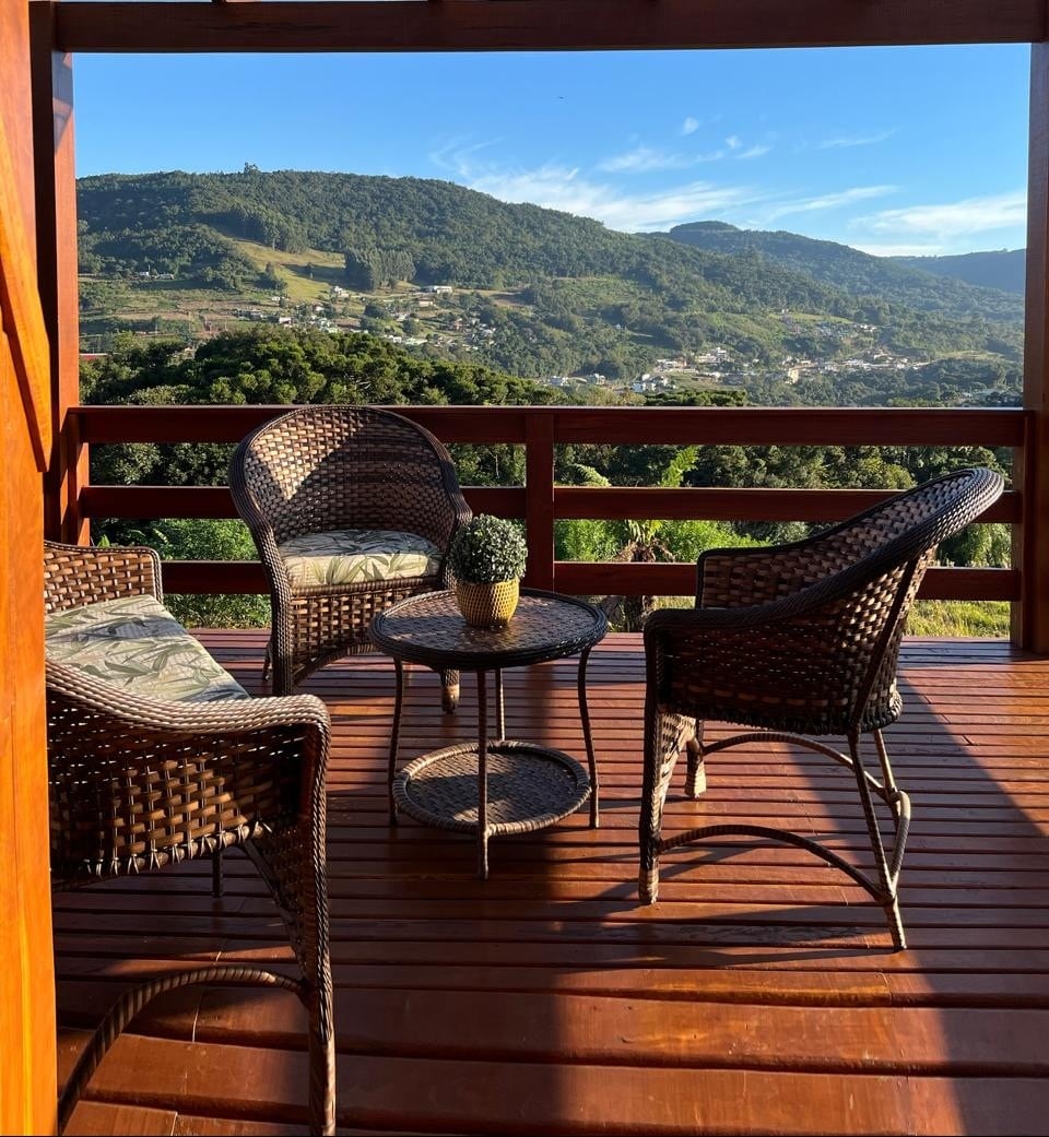 A shaded deck area is furnished with four wicker chairs arranged around a small round table. The surrounding wooden railing offers an unobstructed view of the lush green mountains and valleys, enhancing the natural setting.