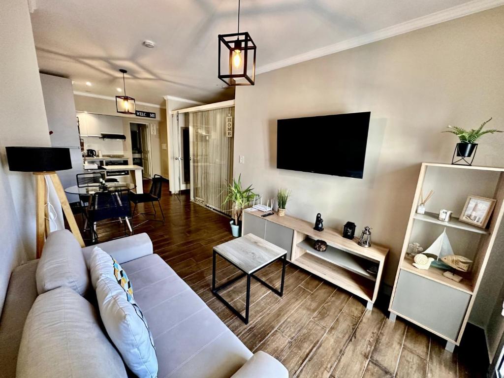 A modern living area is presented with a light grey sofa facing a wall-mounted television. A wooden coffee table sits in front of the sofa, and a stylish shelving unit displays small decorative items. Natural light filters in from a nearby window, creating a bright space.