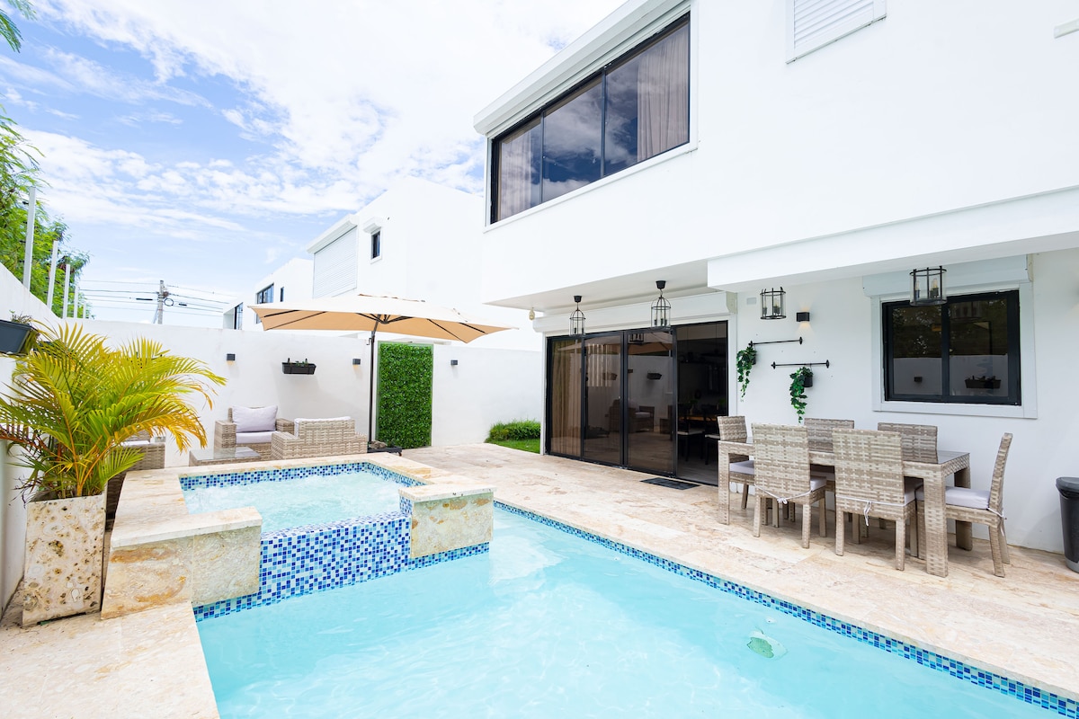 An outdoor area features a tranquil pool, with a separate jacuzzi and elegant tile accents. Comfortable furniture, including a dining set and sun umbrella, are positioned for outdoor relaxation. Large windows offer views into the villa, and greenery enhances the ambiance.