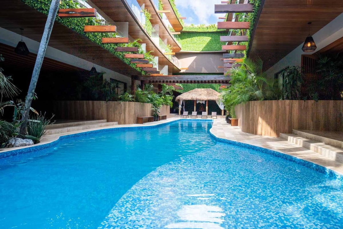 A large, inviting swimming pool is surrounded by lush greenery, featuring wooden decking and lounge chairs. The pool area is illuminated with natural light, creating a serene atmosphere. A thatched-roof hut is visible at the far end, offering a shaded relaxation space.