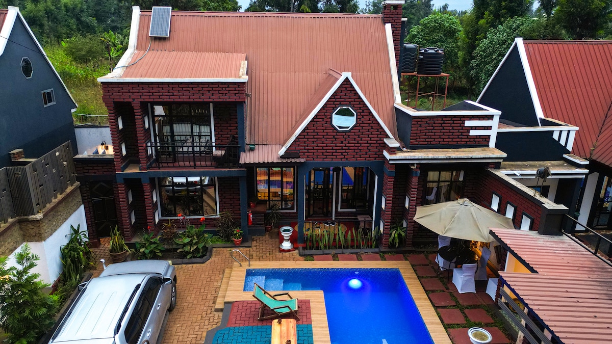 The two-story house, constructed with a mix of red brick and dark siding, is complemented by a swimming pool that reflects bright blue water. Outdoor seating areas and a shaded umbrella are arranged beside the pool, while greenery borders the property.