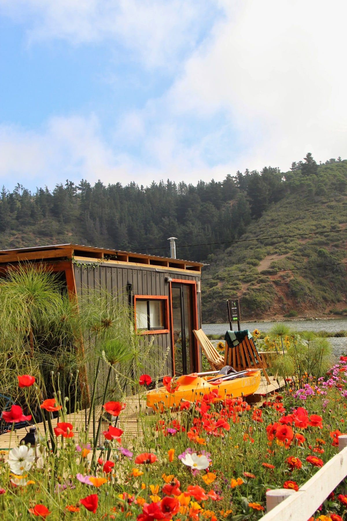 A tiny house sits amidst a vibrant flower garden, featuring an array of colorful blooms. Kayaks are positioned nearby, with a wooden deck leading to the water. Lush greenery and rolling hills rise in the background under a partly cloudy sky.