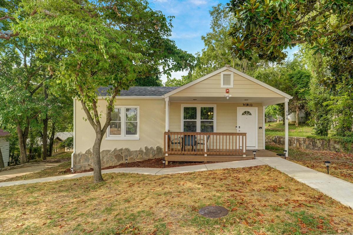 A charming cottage exterior features a light-colored facade and a welcoming front porch with wooden railing. Greenery surrounds the home, including trees and lawn, while a paved walkway leads to the entrance. The roof is gabled and complemented by a small front window.