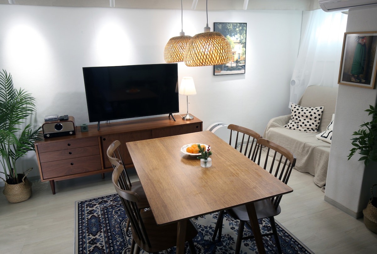 A comfortable living space features a large TV mounted on the wall above a wooden media console. A dining table with six chairs sits in the center, holding a fruit plate. Cozy seating with decorative pillows is arranged nearby alongside a plant for a natural touch.