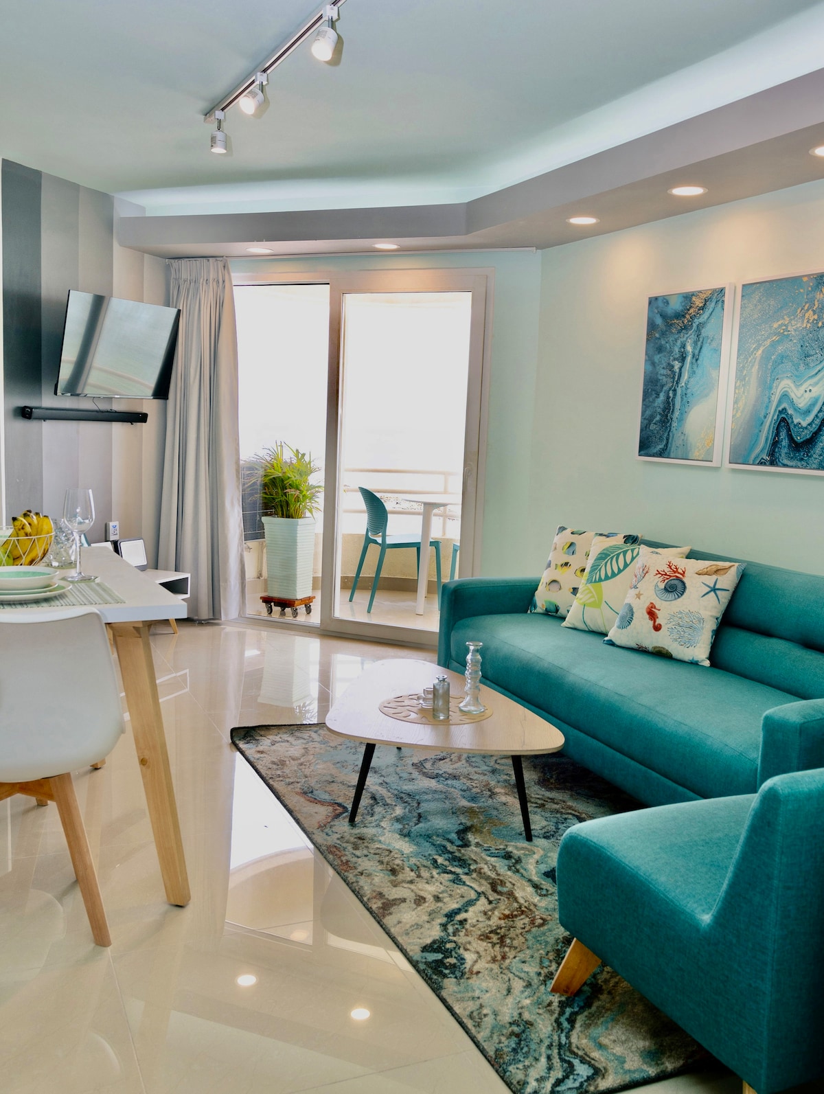 A modern living area is furnished with a comfortable turquoise sofa and a light wooden coffee table, positioned over a patterned rug. A flat-screen TV is mounted on the wall. Natural light enters from the balcony, where greenery is visible. Artistic wall art complements the decor.