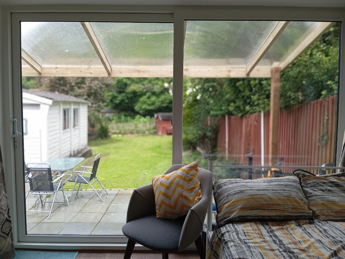 A view from the studio showcases a well-maintained garden, featuring green grass and a patio with outdoor furniture. A cozy chair with patterned cushions is positioned near the glass doors, providing a bright and inviting atmosphere with natural light streaming in.