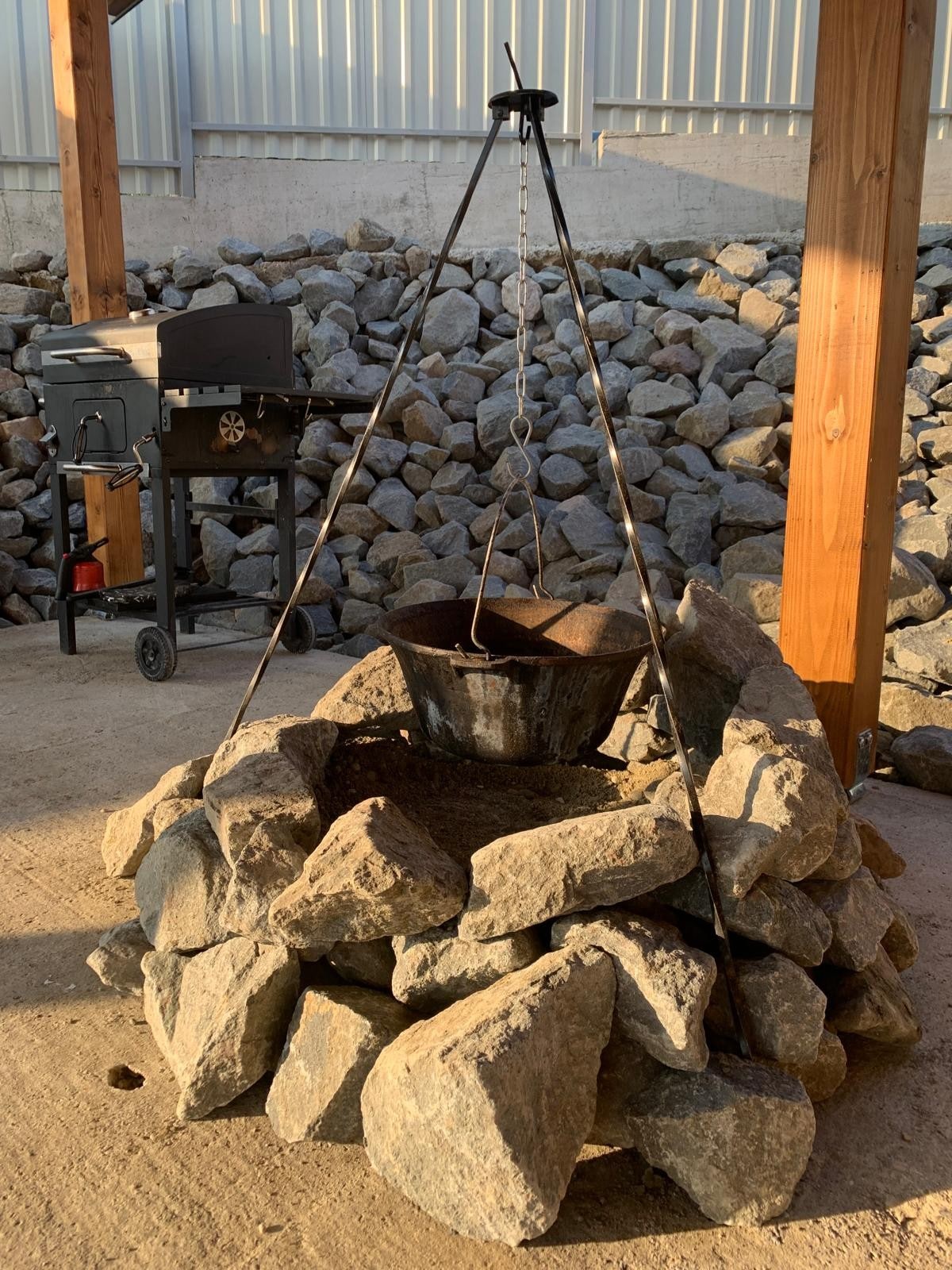 A cooking area is set up with a stone fire pit, featuring a tripod structure and a hanging black pot. Gravel and larger rocks surround the base, providing stability. In the background, a grill is positioned against a wall lined with gravel, creating a rustic outdoor cooking space.