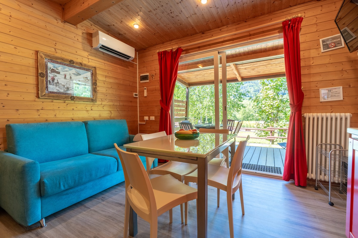 An inviting living area features a turquoise couch and a dining table surrounded by white chairs. Large sliding doors open to a deck, allowing natural light to fill the space. Red curtains frame the windows, adding a touch of color to the wooden walls.