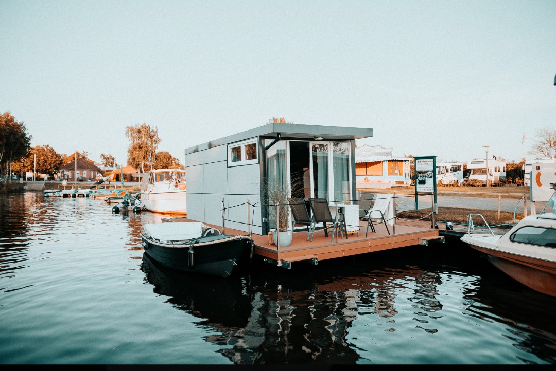 A modern houseboat is anchored at the harbor, surrounded by calm waters. It features a spacious deck with seating arrangements. Nearby, various boats are visible, and the landscape includes lush greenery and structures along the shore.
