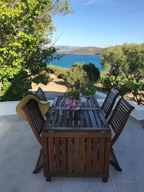 A wooden dining table is set on a tiled patio, surrounded by chairs. A small bouquet of flowers sits in the center, with a sun hat resting on the table. The ocean and lush greenery can be seen in the background under a clear sky.