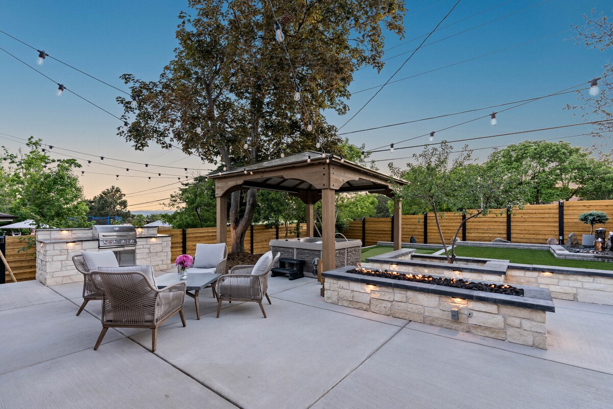 An outdoor seating area features a shaded pavilion, surrounded by trees and greenery. Seating is arranged near a stone fire pit with a gas flame, and an outdoor kitchen is visible. Soft string lights create a warm ambiance above the space.
