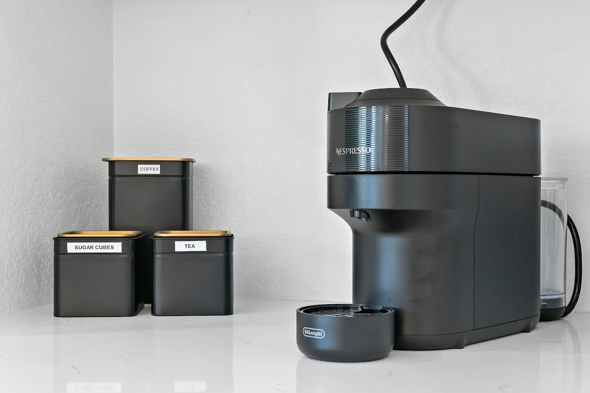 A sleek Nespresso machine is elegantly positioned on a clean, minimalist countertop. Nearby, three labeled canisters contain coffee, sugar cubes, and tea, all featuring contrasting gold lids, enhancing the kitchen's modern aesthetic.