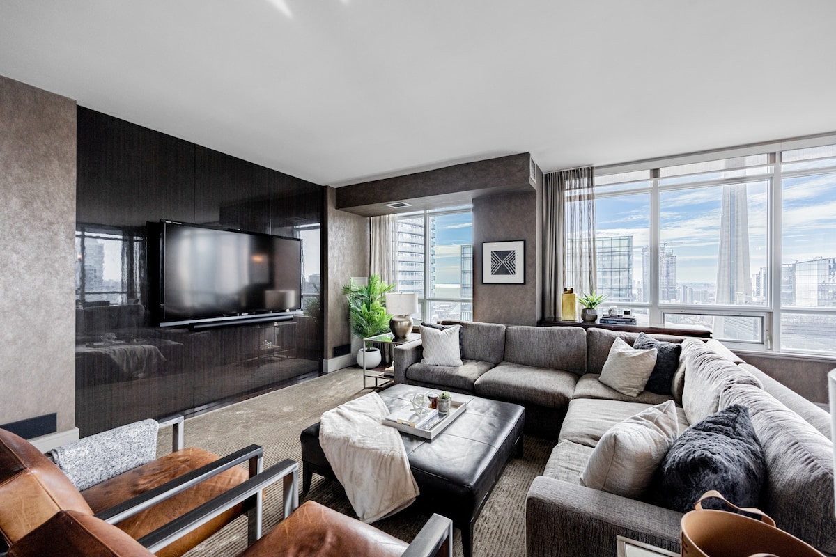 A spacious living area features a large sectional sofa arranged around a coffee table, with sleek furniture and a wall-mounted HD TV. Floor-to-ceiling windows illuminate the room, presenting a panoramic view of the city skyline. Decorative plants add a touch of greenery.