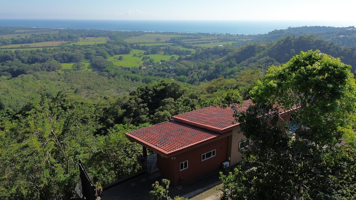 An aerial view of the villa showcases its red-tiled roof positioned amidst lush greenery. The Pacific Ocean glistens in the background, providing a striking contrast to the surrounding landscapes of hills and valleys.
