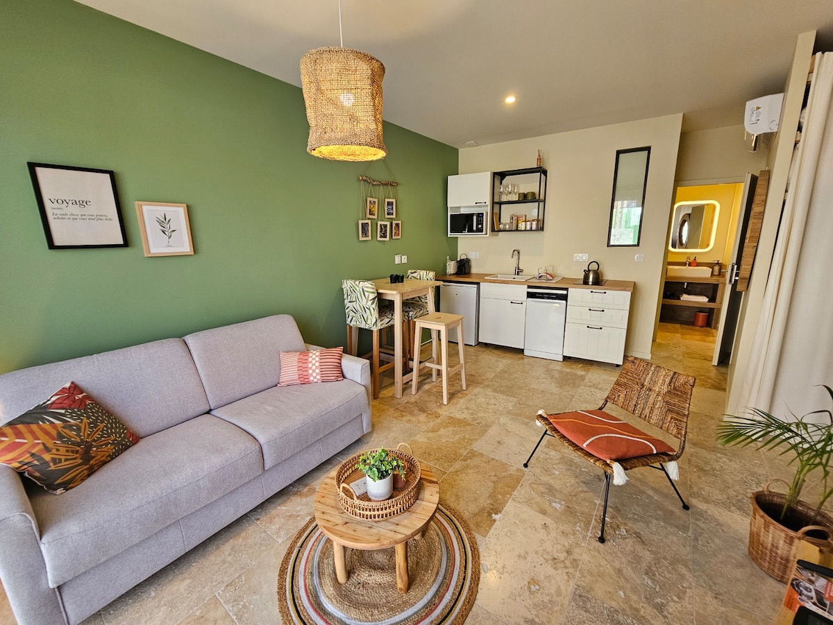 A welcoming living area features a light gray sofa with decorative pillows, complemented by a round coffee table. The kitchen is equipped with modern appliances against a green accent wall. A dining area with wooden stools provides a casual setting. Natural light illuminates the space.