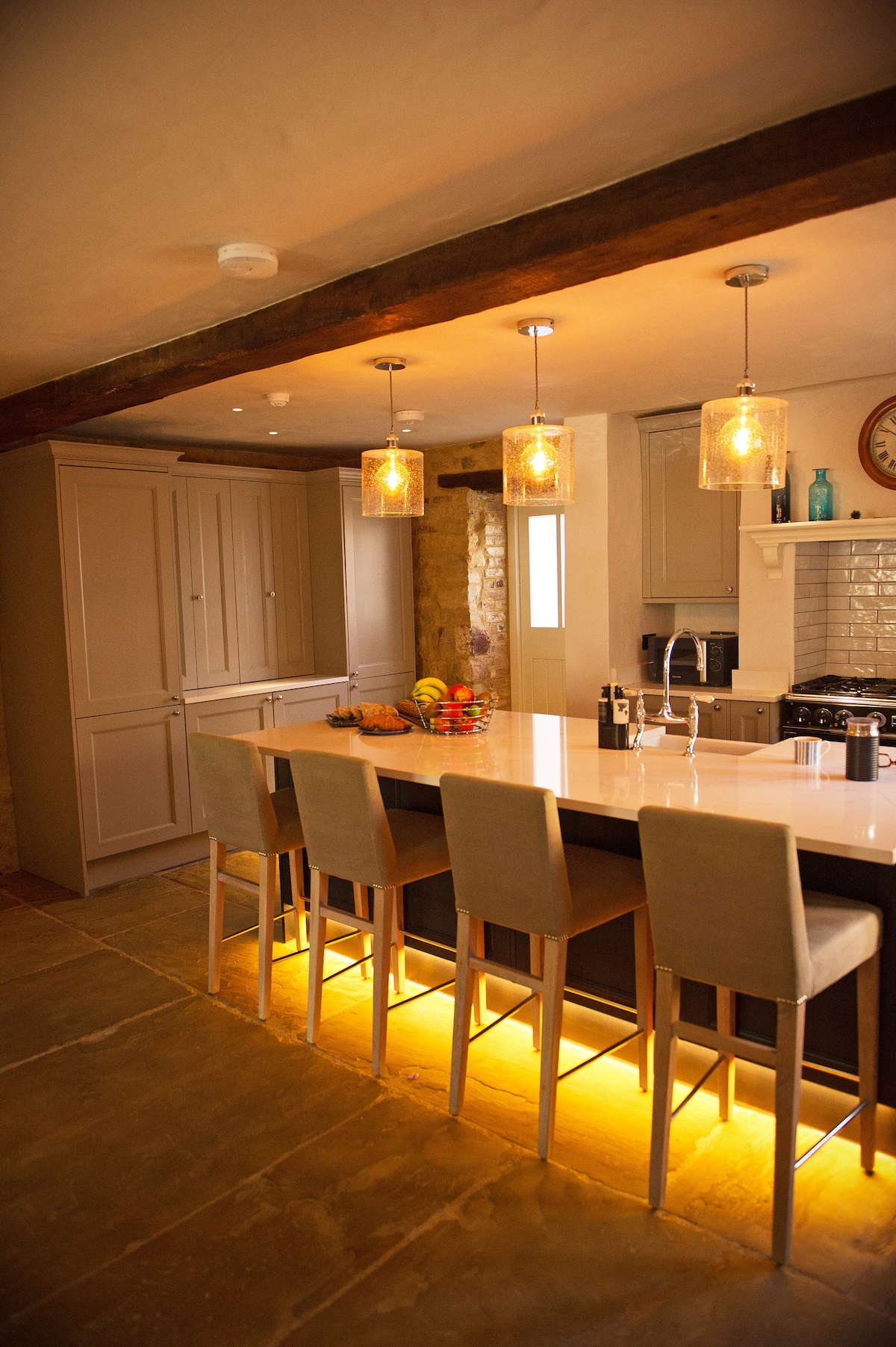 A modern kitchen features sleek cabinetry and a spacious island with seating for four. Soft lighting illuminates the area, enhancing the warm tones of the stone floor and rustic beams above. A subtle arrangement of fruits is displayed on the island.