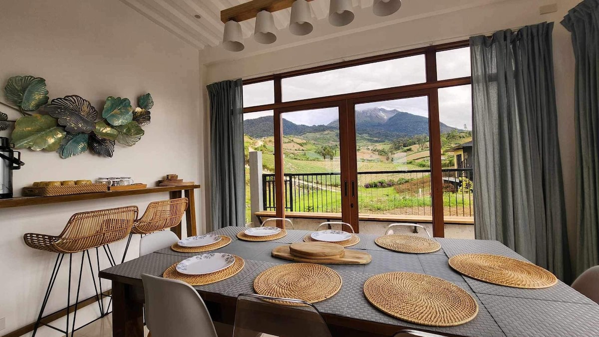A dining area features a wooden table set with woven placemats and plates, surrounded by stylish chairs. Large glass doors open to a scenic view of rolling hills and Mt. Apo. Soft curtains frame the windows, allowing natural light to brighten the space.