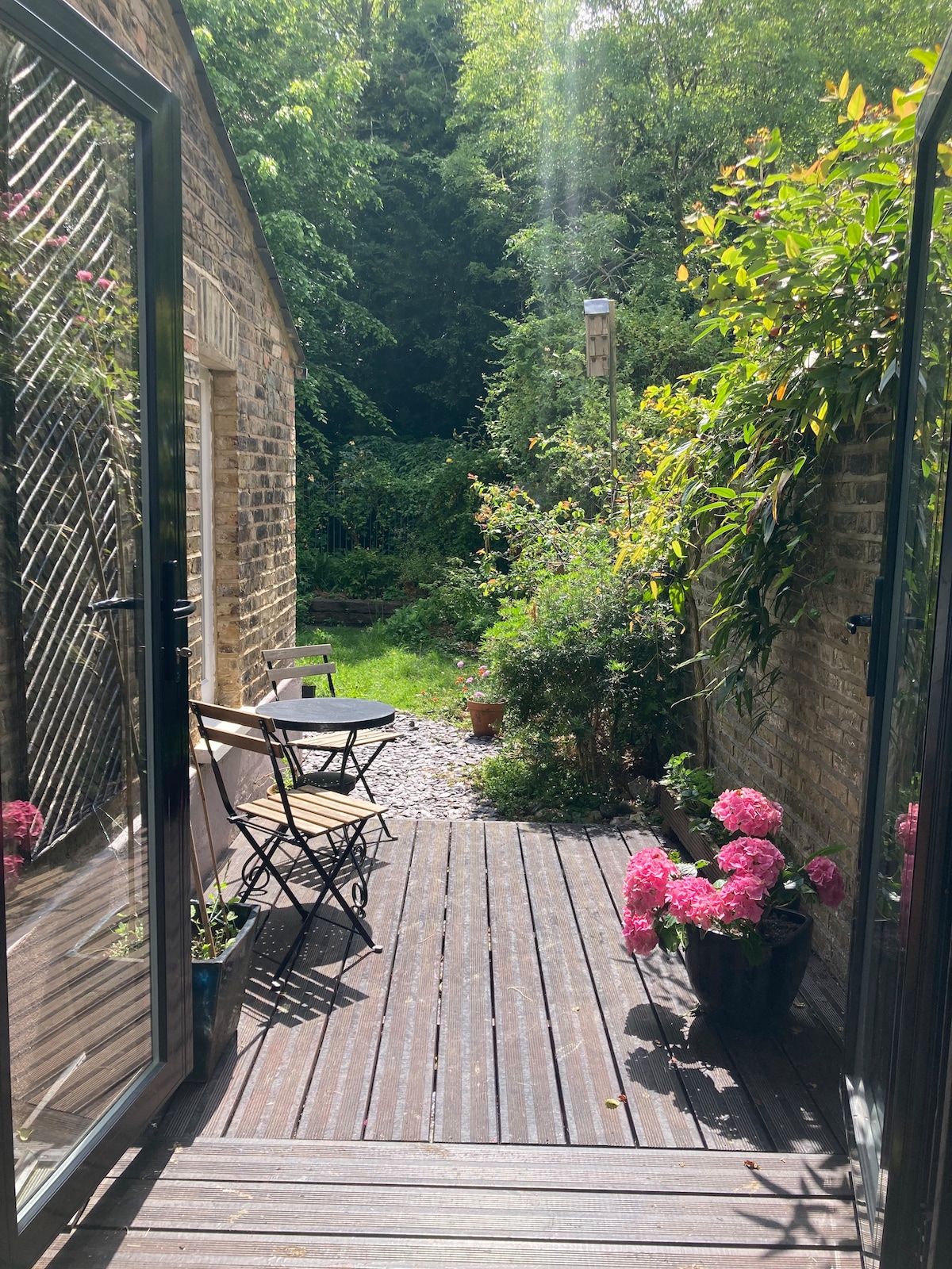 A wooden deck area is framed by glass doors, leading to a garden adorned with vibrant pink flowers in pots. A small bistro set is visible, inviting relaxation in a serene outdoor space surrounded by greenery.