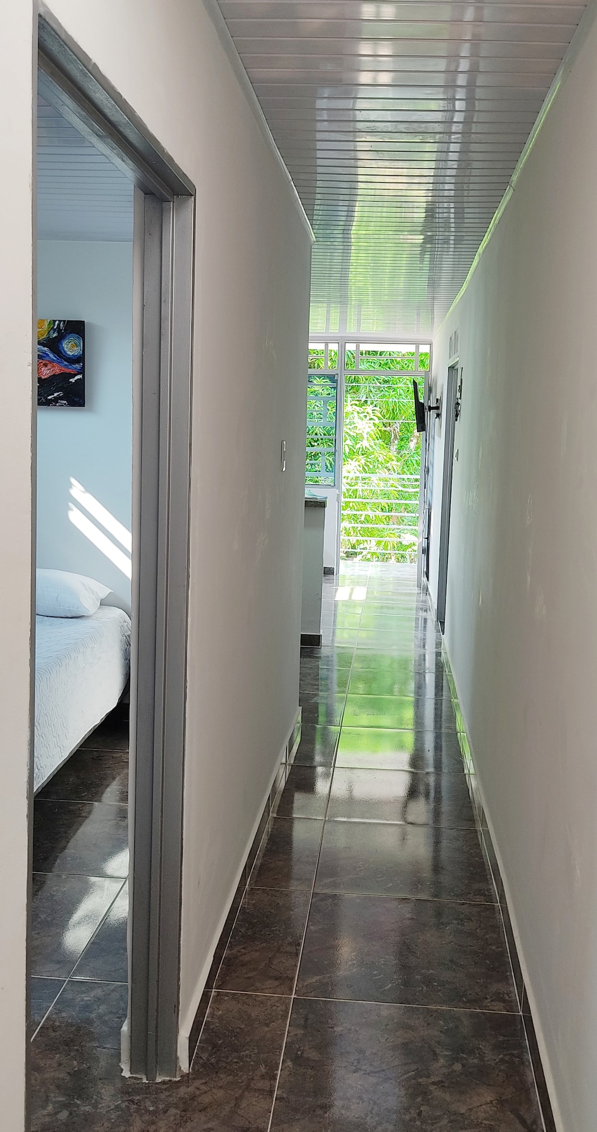 A well-lit hallway leads to a spacious room, with natural light filtering through large windows. The tiled floor reflects the light, creating a bright and inviting passage. A glimpse of greenery is seen through the window, enhancing the connection with nature.
