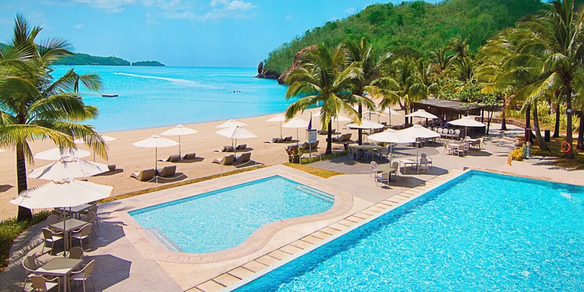 The image depicts a serene beach setting featuring two swimming pools adjacent to the sandy shoreline. Umbrella-shaded lounge chairs are positioned around the pools, surrounded by tropical palm trees. The calm blue water of the sea meets a clear sky, creating a tranquil atmosphere.