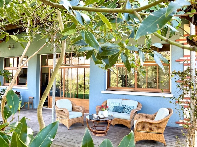 A spacious covered terrace is framed by lush greenery, featuring a cozy seating arrangement with two wicker chairs and a small table. Large windows allow natural light to fill the area, enhancing the inviting outdoor space overlooking the garden.