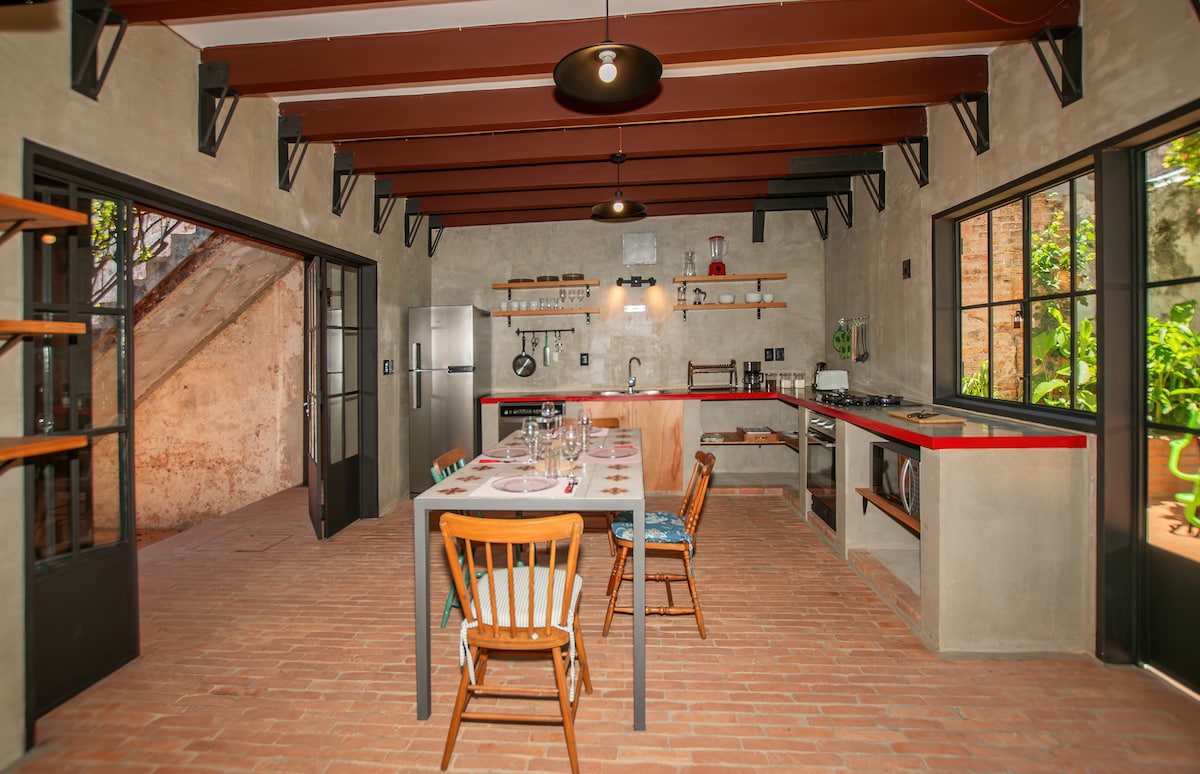 A spacious kitchen features rustic charm with brick flooring and wooden beams. An inviting dining table is set for meals, accompanied by colorful chairs. Modern appliances, including a large refrigerator and gas stove, are complemented by open shelving displaying kitchenware.