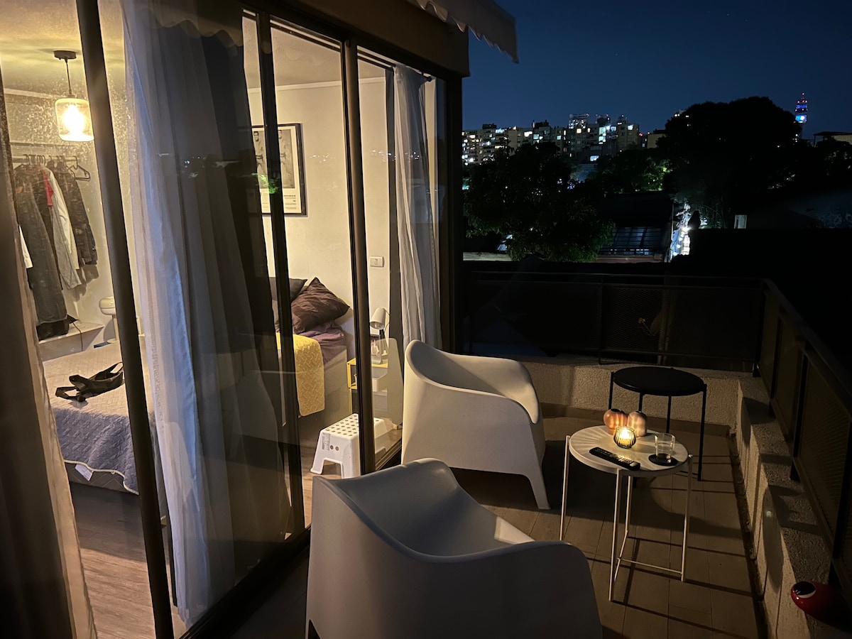 An outdoor balcony area is visible, featuring two modern white chairs and a small table. The space is illuminated softly by candles, with the city skyline of Santiago visible in the background. Light drapes hang alongside a large glass door, leading into a cozy interior.