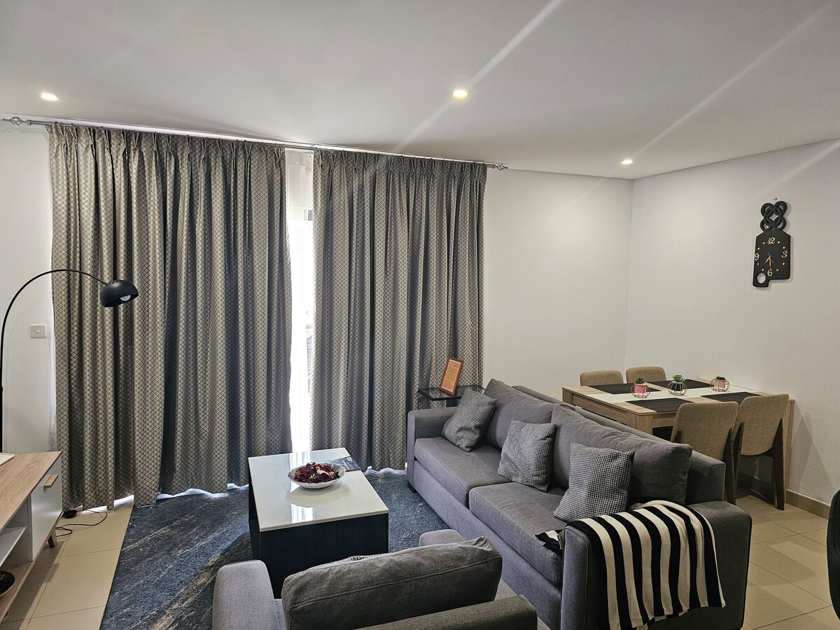 A modern living area is presented with comfortable gray sofas arranged around a central coffee table. Large curtain panels frame the windows, allowing soft light to enter. A simple dining table with chairs is visible in the background, enhancing the functional layout.