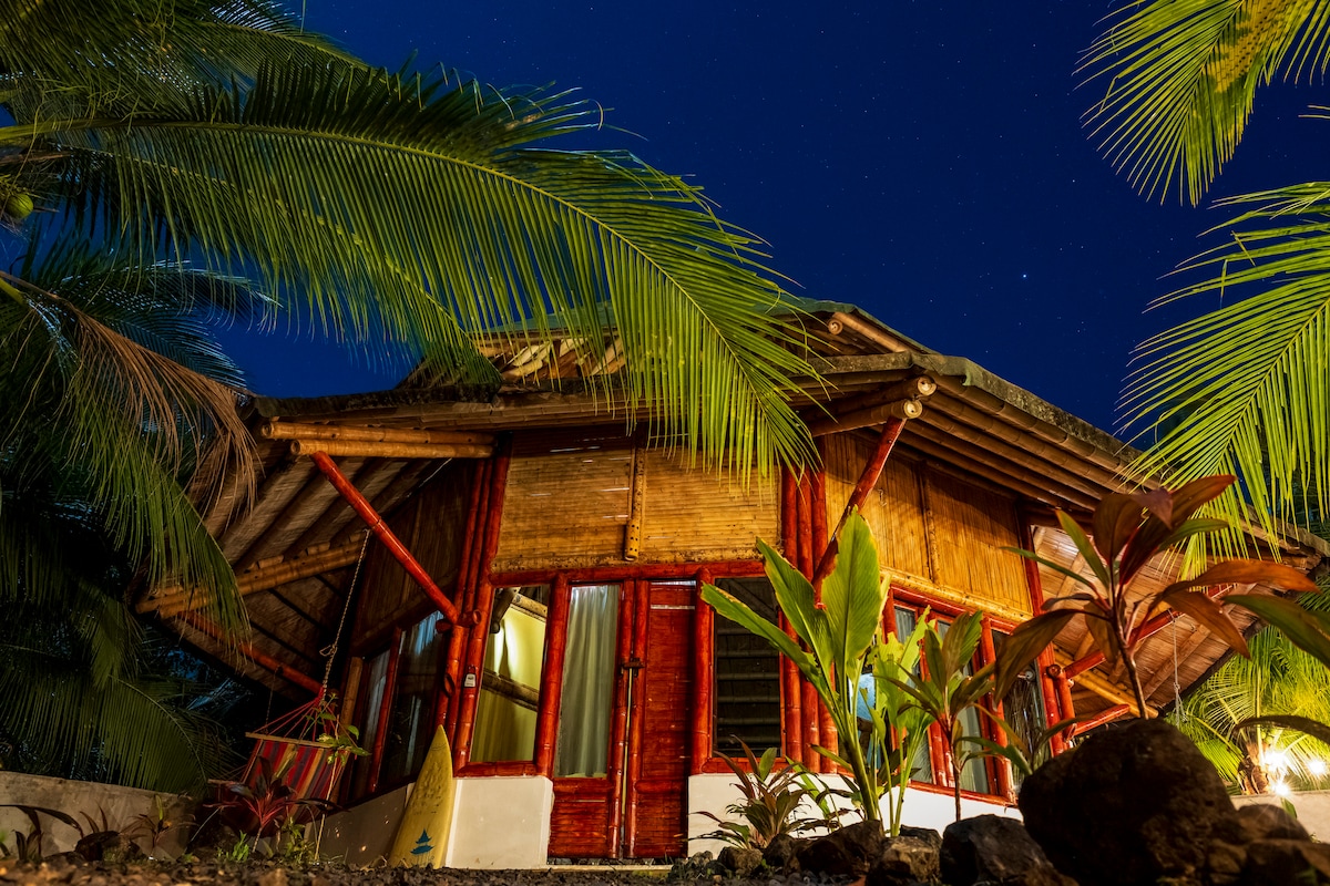 A bamboo cabin is framed by lush palm trees under a clear night sky. The structure features large windows and red wooden accents, showcasing a harmonious blend with nature. Soft lighting provides a warm glow, inviting a sense of tranquility and peace.