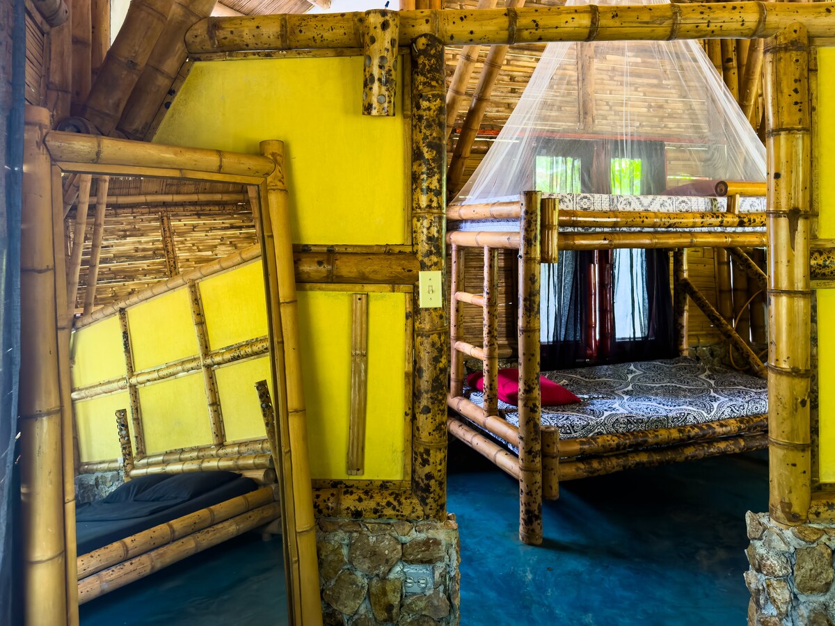 An interior view showcases a cozy sleeping area with a double-decker bamboo bed featuring a patterned mattress. The yellow walls create a bright ambiance, and natural bamboo beams highlight the craftsmanship. A netting drapes above the top bunk, enhancing the rustic appeal.