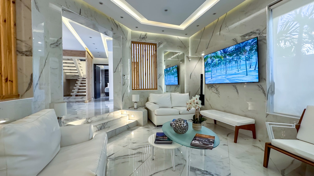 A modern living area is presented with sleek white furniture and marble flooring. A large television is mounted on a wall, while natural light is filtered through the windows. A glass coffee table holds a small potted plant, and a few magazines are neatly arranged beneath it.
