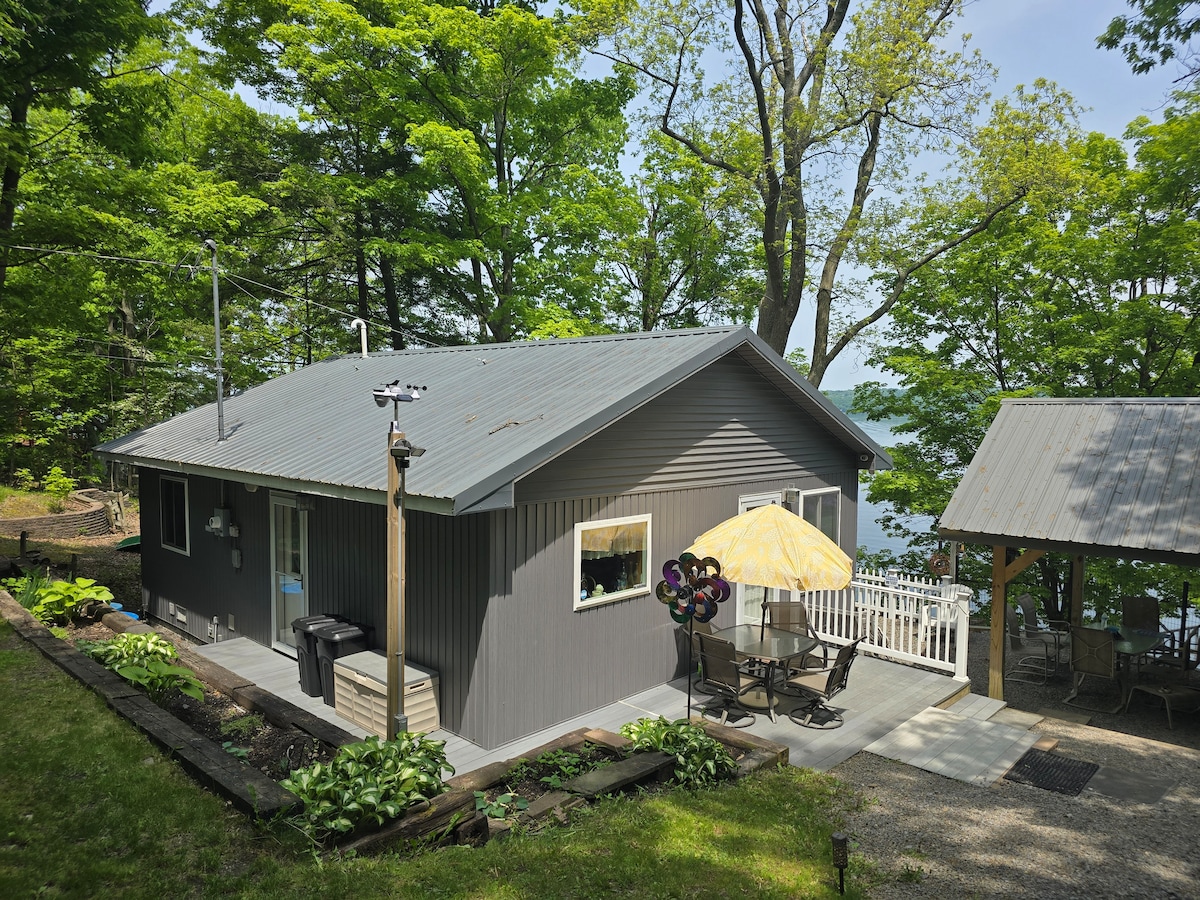 A cozy cottage is nestled among lush green trees, showcasing a neutral exterior with a metal roof. Outdoor patio seating features a yellow umbrella, providing shade. A charming pavilion is visible nearby, and the serene landscape offers a calming backdrop.