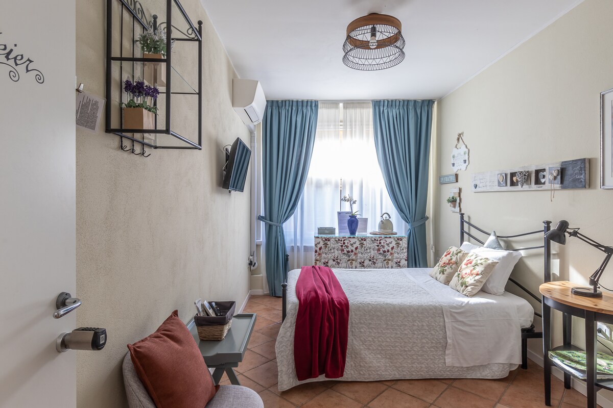 The room features a spacious bed adorned with decorative pillows and a colorful throw. A small television is mounted on the wall, and a wooden table, alongside a comfortable chair, complements the space. Large windows are dressed with flowing blue curtains, allowing natural light to fill the room.