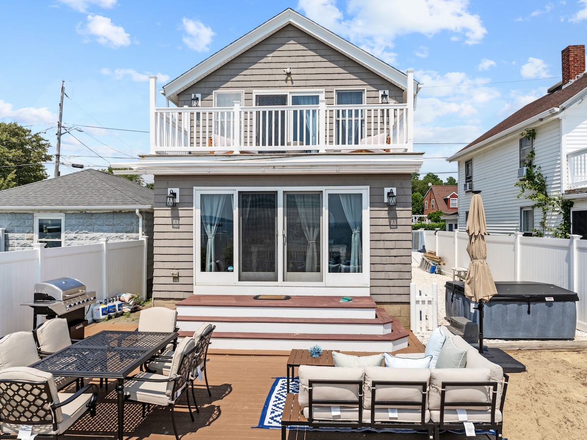 The exterior of the residence features a multi-level structure with a spacious deck on the second floor. Glass doors provide views of the outdoor seating area, which includes a dining set and comfortable lounge furniture. A hot tub and a sandy area are visible, surrounded by a fenced backyard.