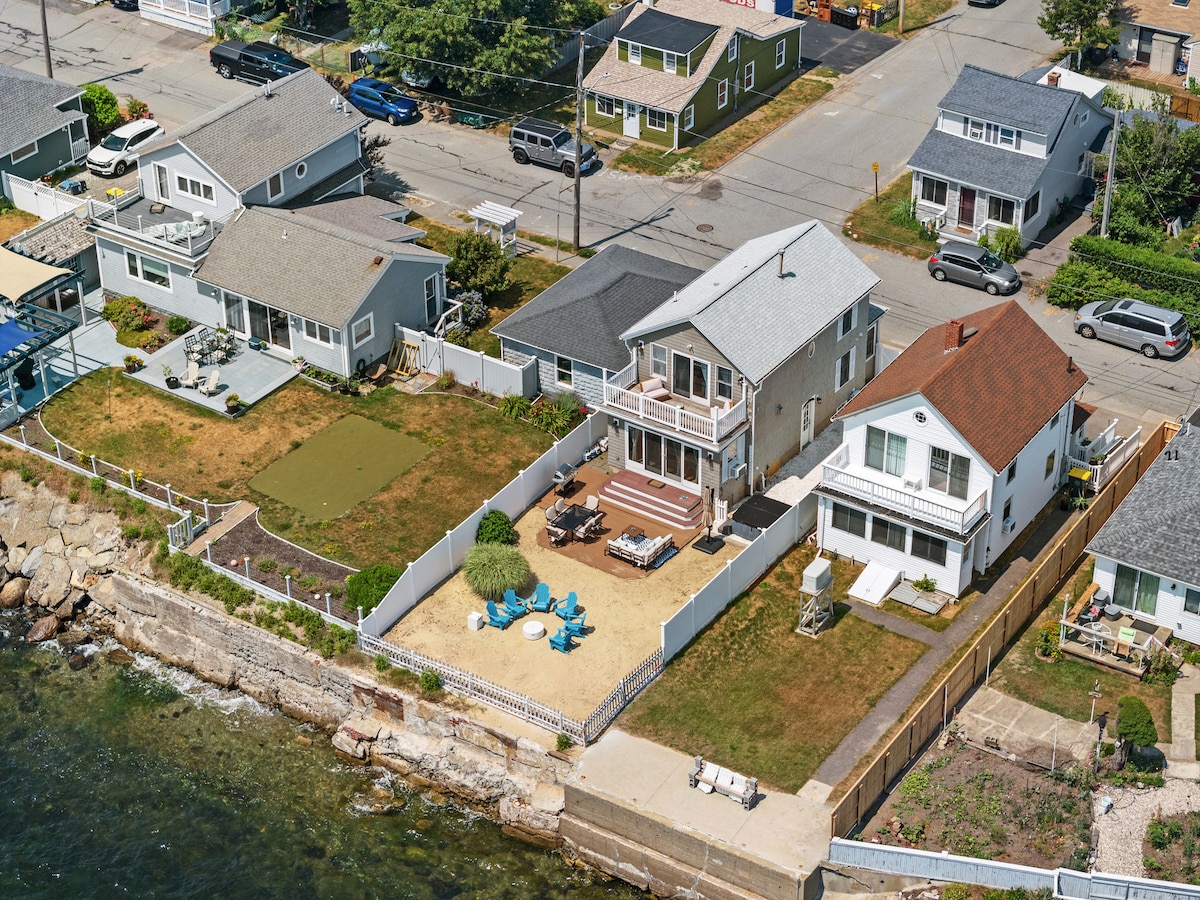 An aerial view showcases a coastal property with a sandy backyard and a fenced area. Several outdoor seating arrangements are visible, including chairs and lounge areas. Nearby houses and streets line the shoreline, which gently meets the water, highlighting its waterfront location.