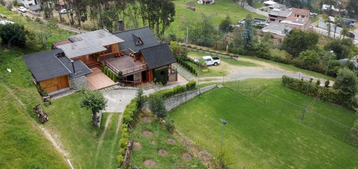 The property is captured from an aerial view, showcasing the spacious grounds and surrounding natural landscape. The main building, featuring multiple rooflines and a large patio, is accompanied by open green areas, a sports field, and well-manicured gardens.