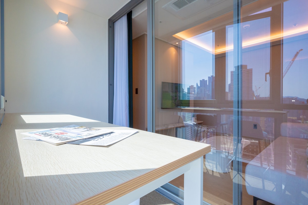 A bright indoor area features a modern table with magazines spread across its surface. Large glass doors provide a clear view of the outdoor cityscape and allow natural light to fill the space, accentuating the clean lines and contemporary design.