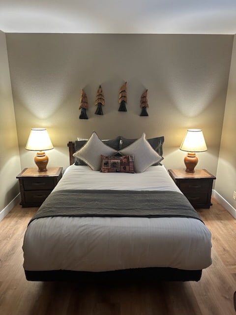 A welcoming bedroom displays a spacious bed with neutral bedding and decorative pillows. Nightstands with lamps flank each side, providing soft illumination. The walls are adorned with wooden wall art, enhancing the room's cozy atmosphere.