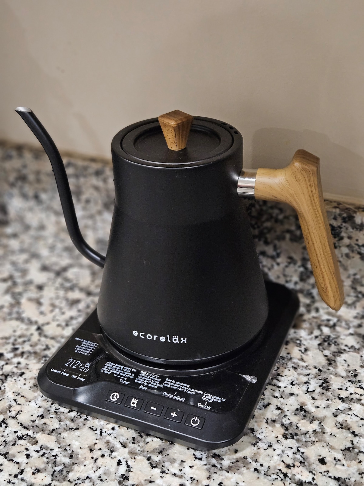 A sleek, black kettle with a wooden handle sits on a countertop. The kettle features a spout for precise pouring and is located atop an electric base with control buttons. The surface is speckled gray, enhancing the modern aesthetic.
