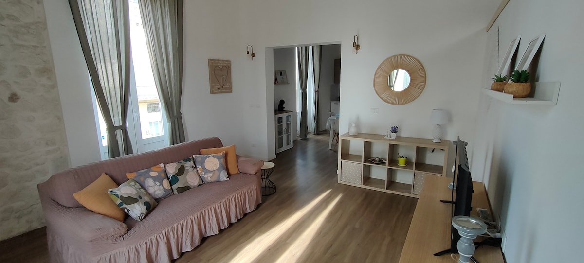 A bright living area features a soft pink sofa adorned with patterned cushions, complemented by natural light streaming through large windows. A wooden console showcases decorative items, while a flat-screen TV is positioned nearby on a minimalistic shelf.