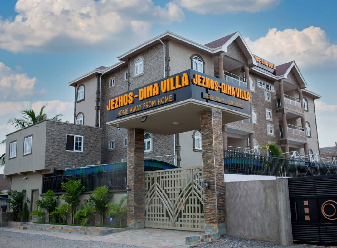The exterior of JezHos-Dina Villa is featured, showcasing a modern building with a welcoming entrance. The stone facade is complemented by vibrant signage stating 'Home Away From Home.' Lush greenery surrounds the entrance, contributing to a serene atmosphere.