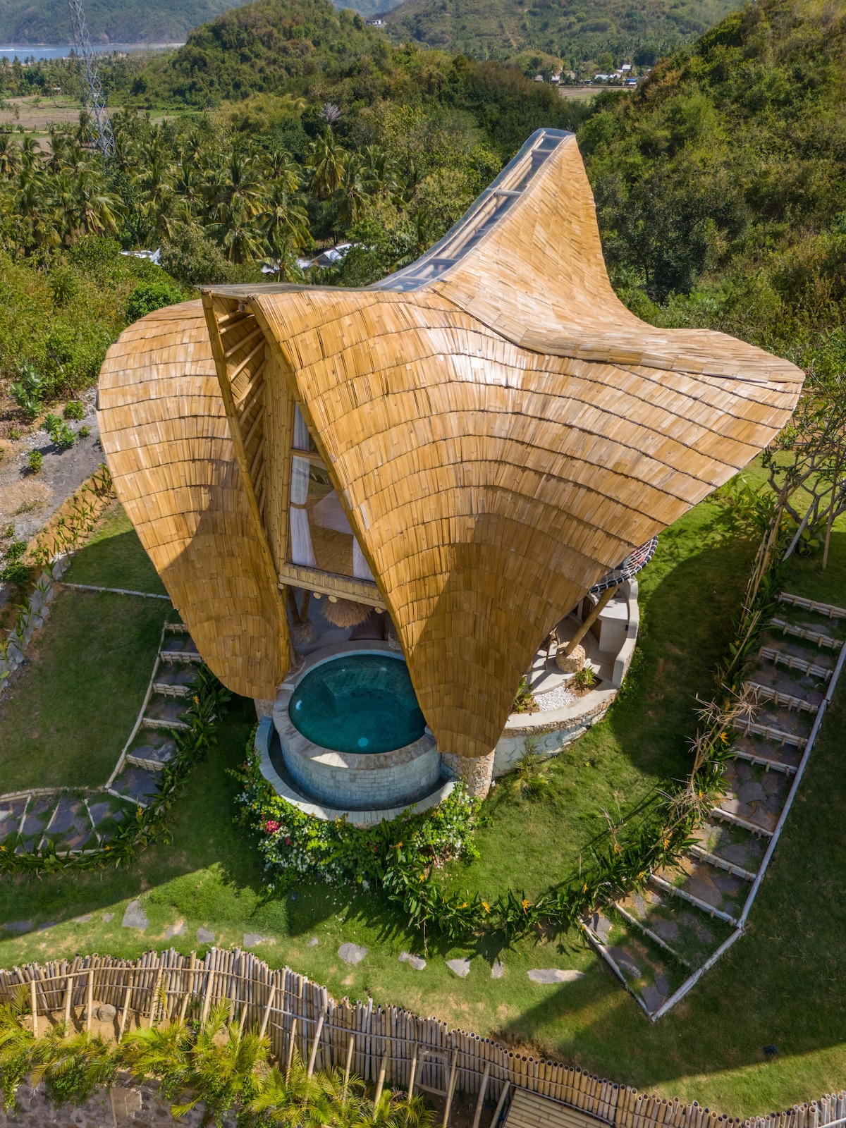 The unique bamboo villa is surrounded by lush greenery, showcasing a distinctive curvilinear design. A private pool is visible on the lower level, complemented by landscaped gardens and winding pathways that enhance the connection with nature.