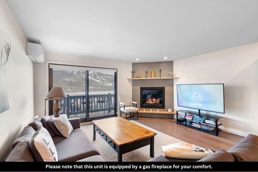A living room area is highlighted by a modern gas fireplace set into a light grey wall. A large television is positioned above a media unit. Comfortable seating includes a couch and armchair, while large glass doors open to a balcony with scenic views.
