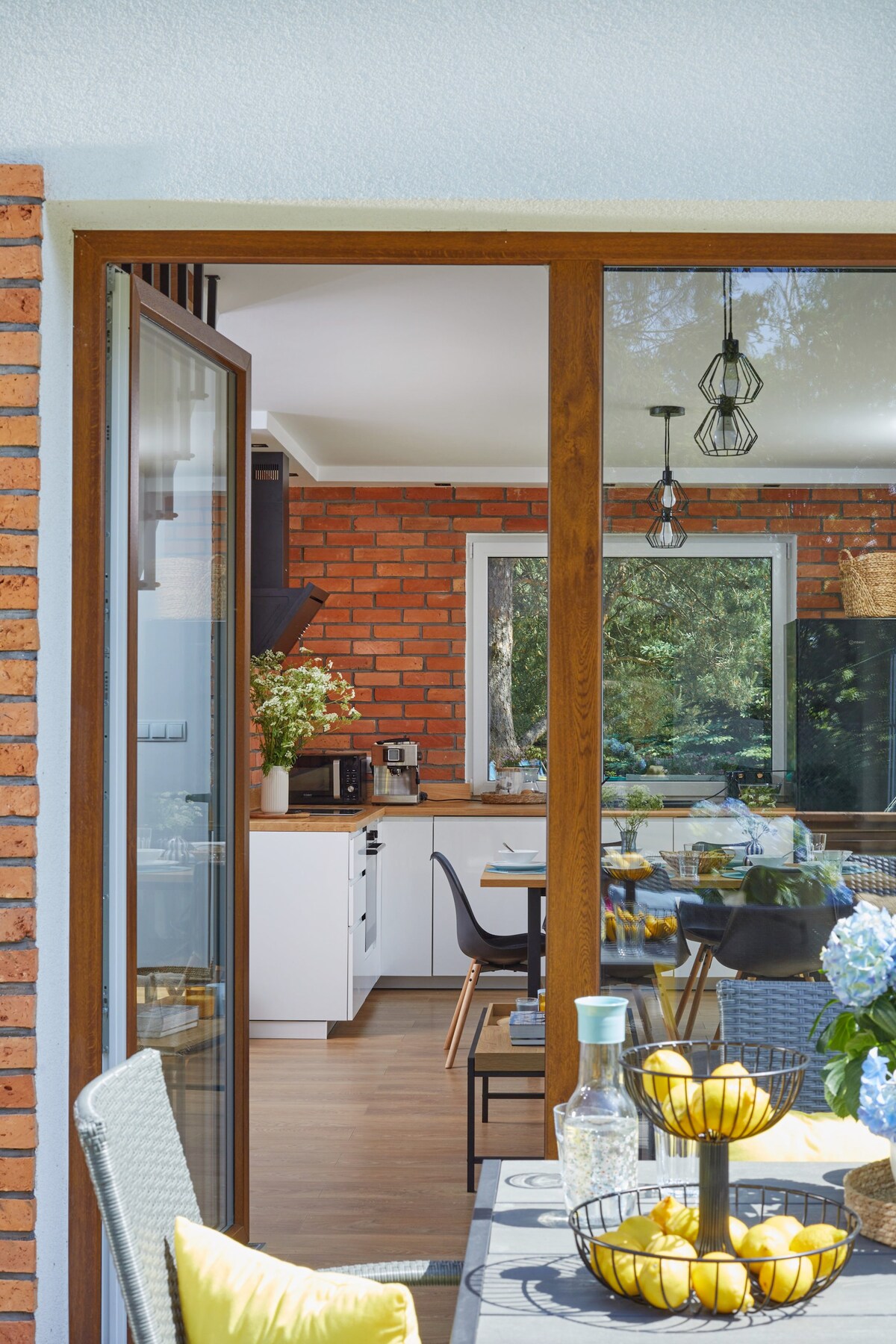 A view from an outdoor terrace showcases a modern kitchen and dining area with wooden accents. The space features a dining table surrounded by chairs, complemented by decorative flowers. Sunshine filters through large windows, illuminating the inviting interior.