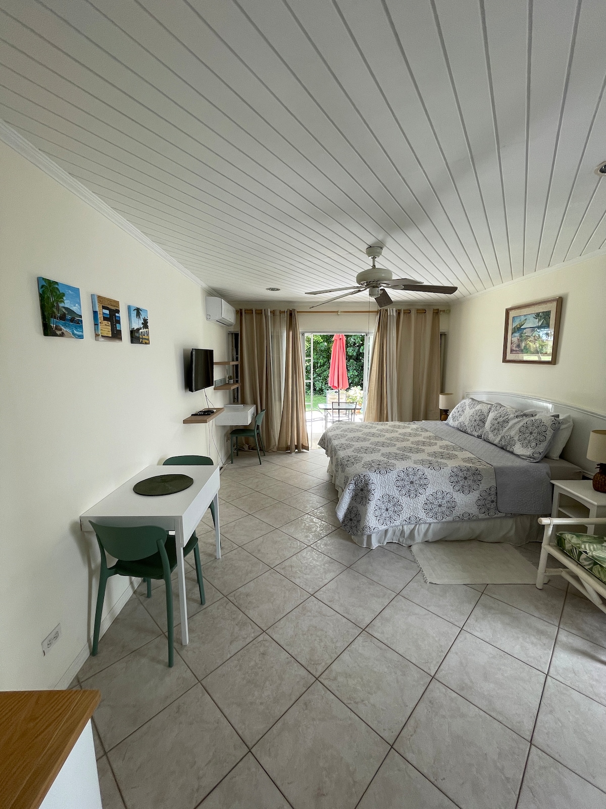 A studio apartment features a spacious bed adorned with a light grey patterned duvet. A small dining table and two chairs are positioned near the window, which has sheer curtains. A flat-screen TV is mounted on the wall, and sliding doors lead to a patio with outdoor seating.