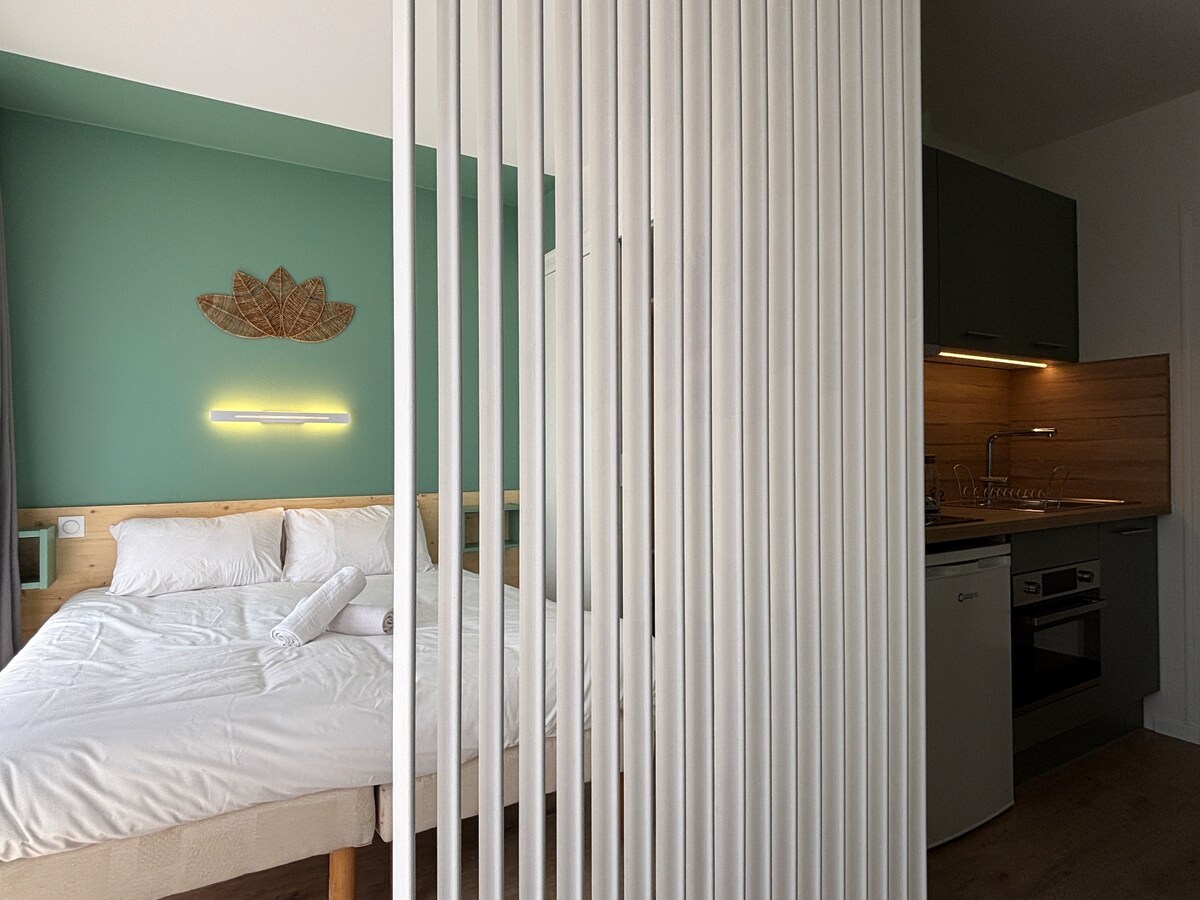A cozy sleeping area is separated by vertical wooden slats, featuring a spacious bed with neatly arranged white linens. A soft green wall provides a calming backdrop, complemented by modern kitchen amenities visible in the background.