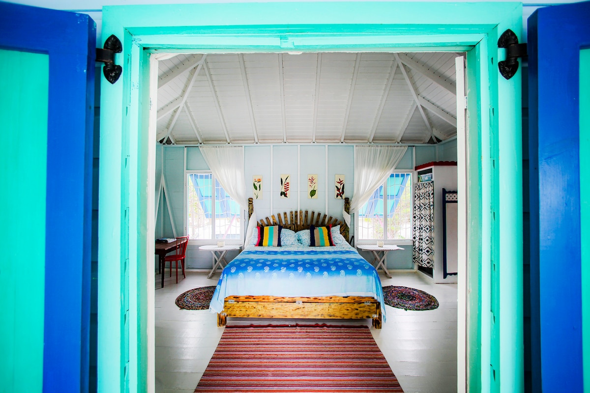 A bright and spacious bedroom is framed by an open doorway. A large bed with a light blue coverlet features colorful throw pillows. Subtle patterns adorn the walls, while natural light flows in through multiple windows, creating a welcoming atmosphere.