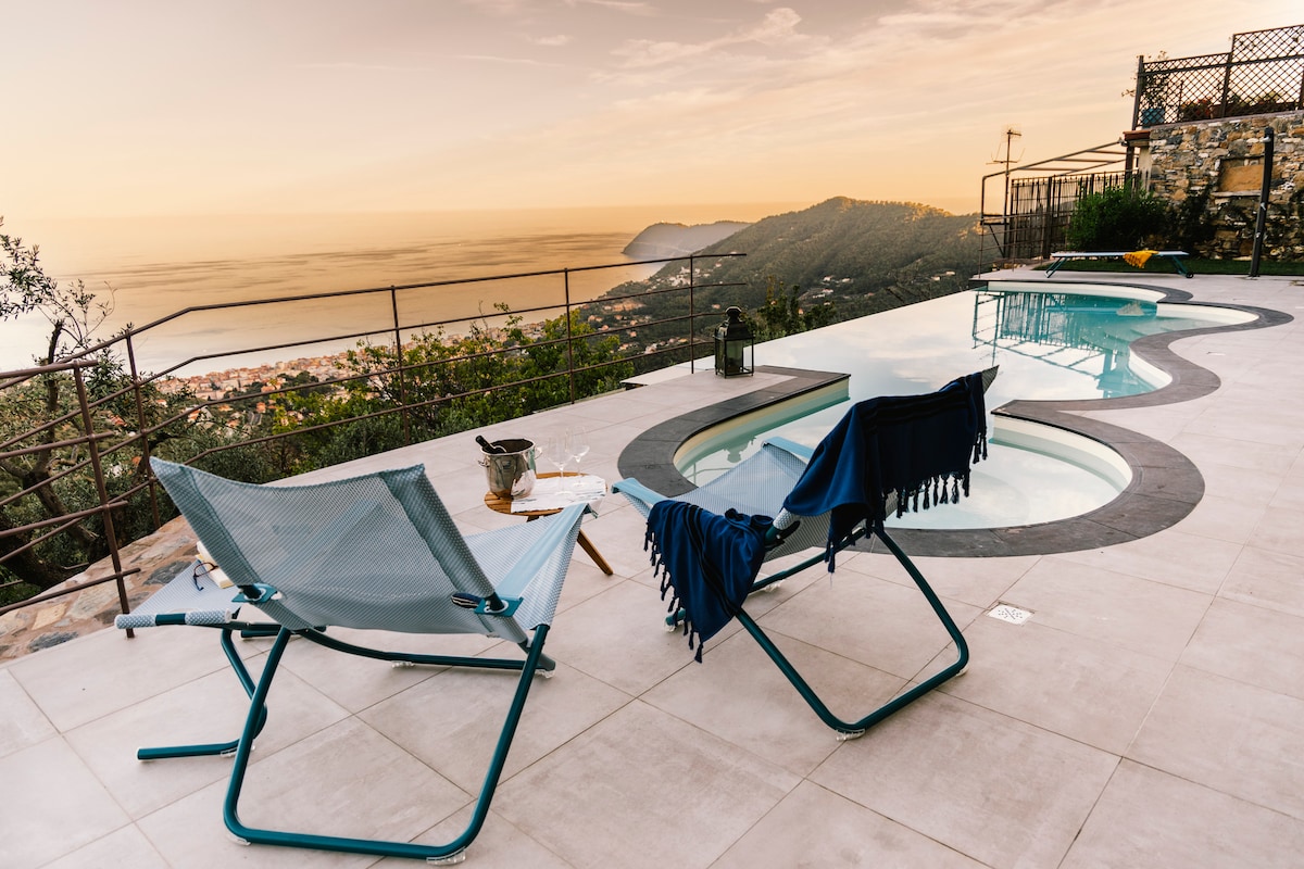 The outdoor area features a uniquely shaped infinity pool beside two blue lounge chairs, one adorned with a folded towel. A small table sits nearby, holding a bottle and glasses. The scene is framed by a panoramic view of the coastline and mountains at sunset.