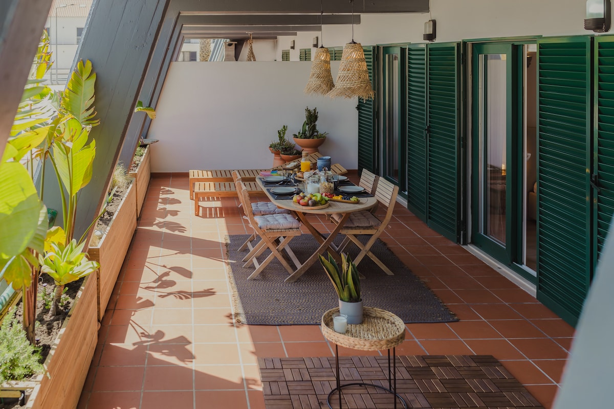 An outdoor dining area is presented with a wooden table set for meals, surrounded by potted plants. The space features wicker pendant lights above and is partially shaded, highlighting the terracotta tiles and wooden accents.
