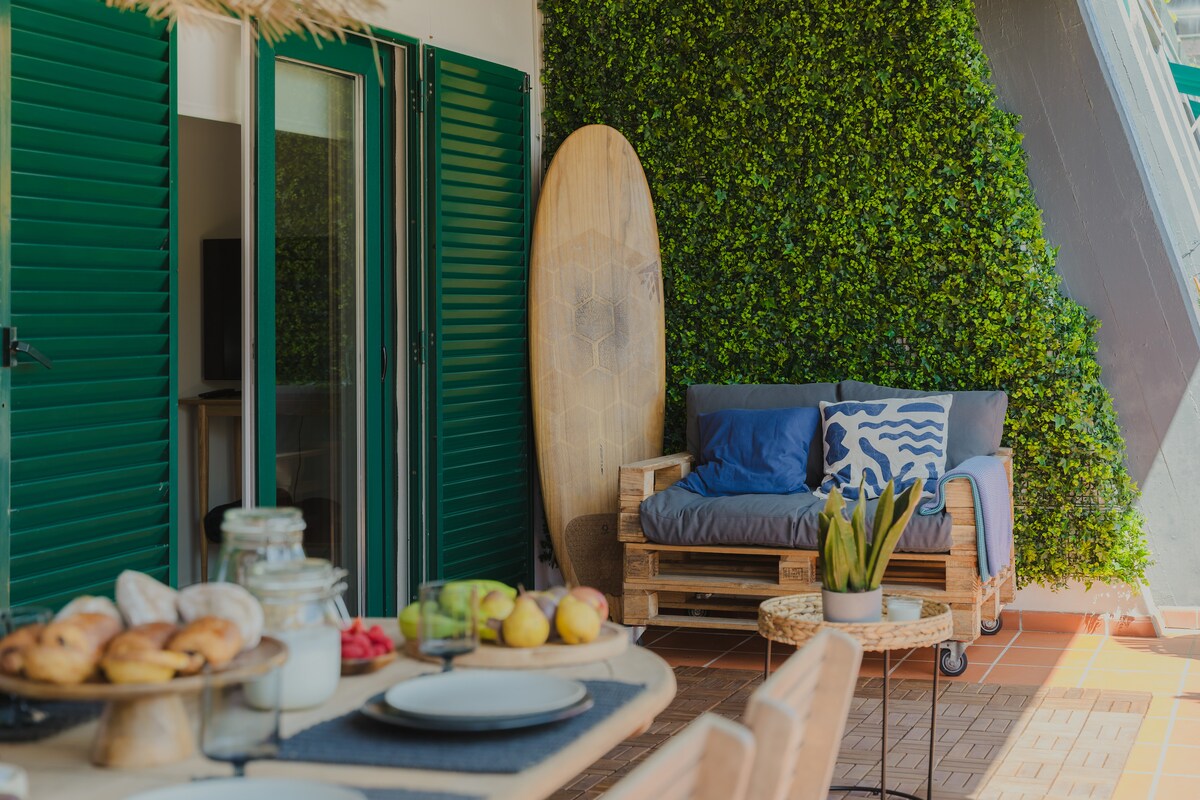 An outdoor terrace area is displayed, featuring a seating arrangement with a grey couch adorned with blue cushions. A wooden surfboard stands against a vibrant green wall. A dining table is set with various fruits, pastries, and a jar, inviting casual dining in the open air.